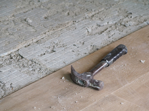 Een hamer ligt op een houten plank, met op de achtergrond een deels verwijderde vloer, met zicht op de ondervloer en wat puin.